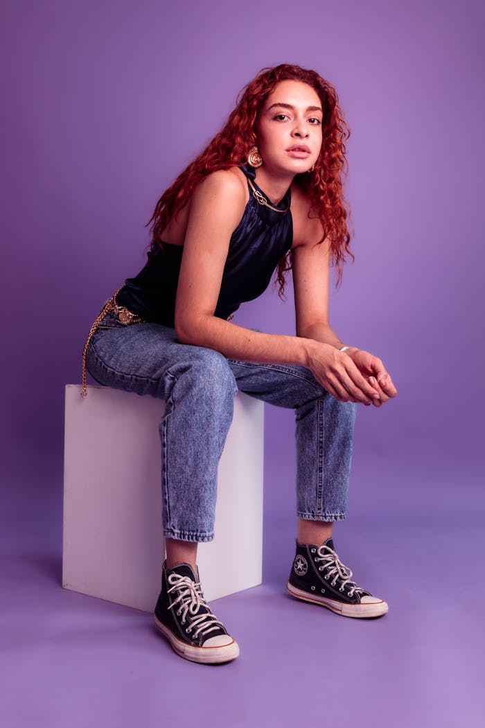 Stylish young woman with red hair and sneakers in a purple-toned studio setting.