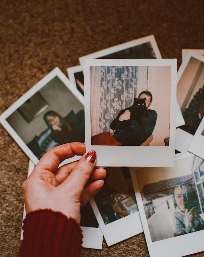 A hand with red nails holds a vintage Polaroid among scattered photos on carpet.